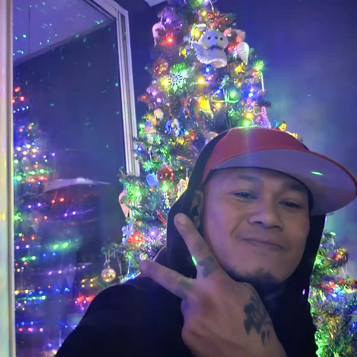 A man wearing a red and white baseball cap and a black hoodie poses for a selfie in front of a brightly lit, colorful…