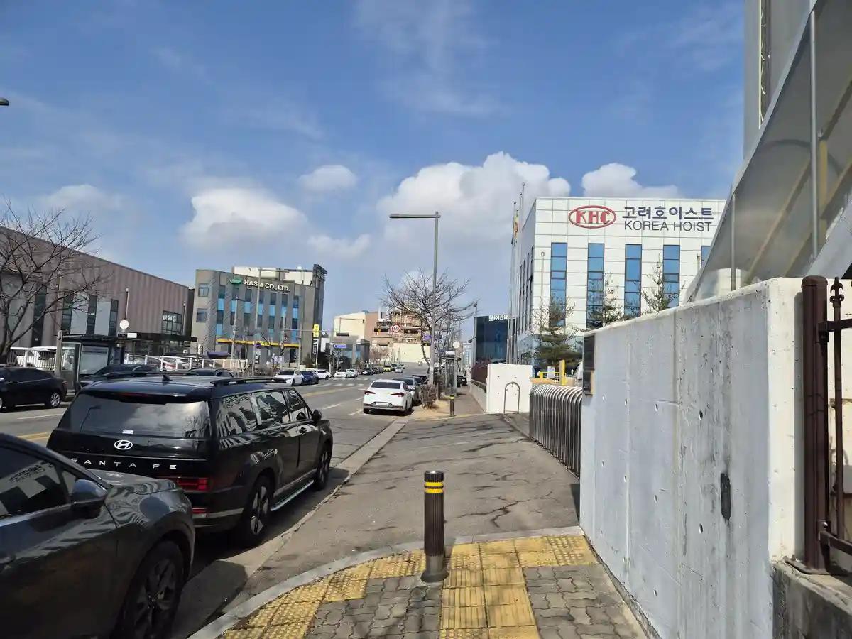 A street view in Korea showing a sidewalk next to a concrete wall and parked cars, with a large building in the background…