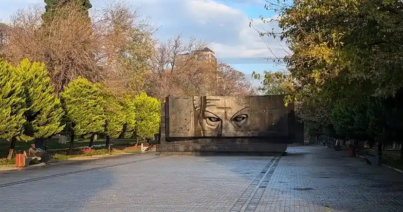 A wide shot of a park pathway leading to a large stone monument with a carved face featuring intense, staring eyes. Trees…