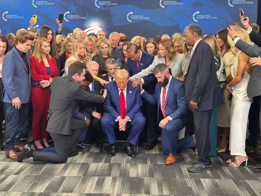a man in a blue suit and red tie sits in a chair surrounded by a crowd of people, some kneeling and placing hands on his…