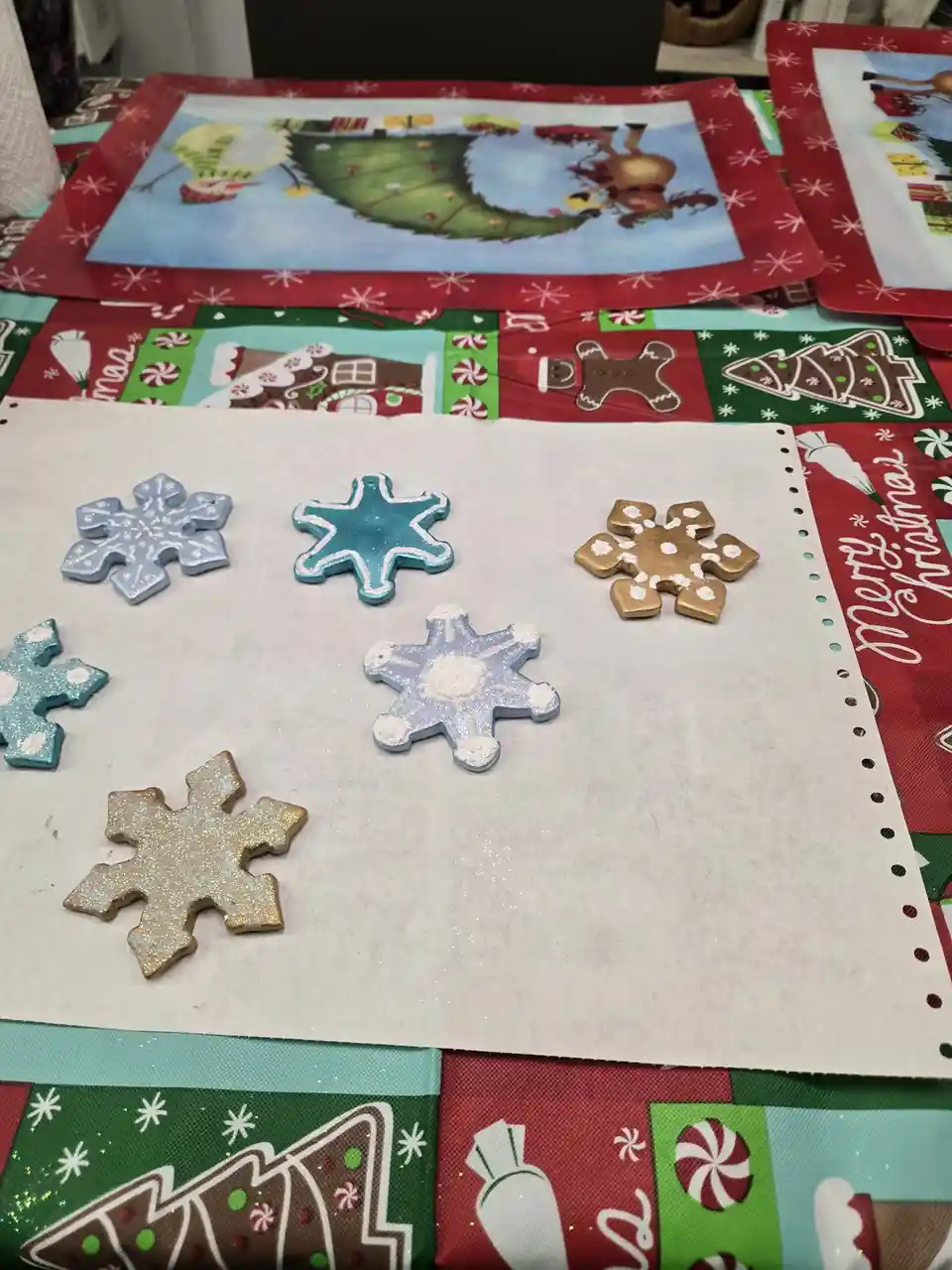 A photo shows several hand-painted, glittery snowflake ornaments on a white sheet of paper, placed on a colorful…