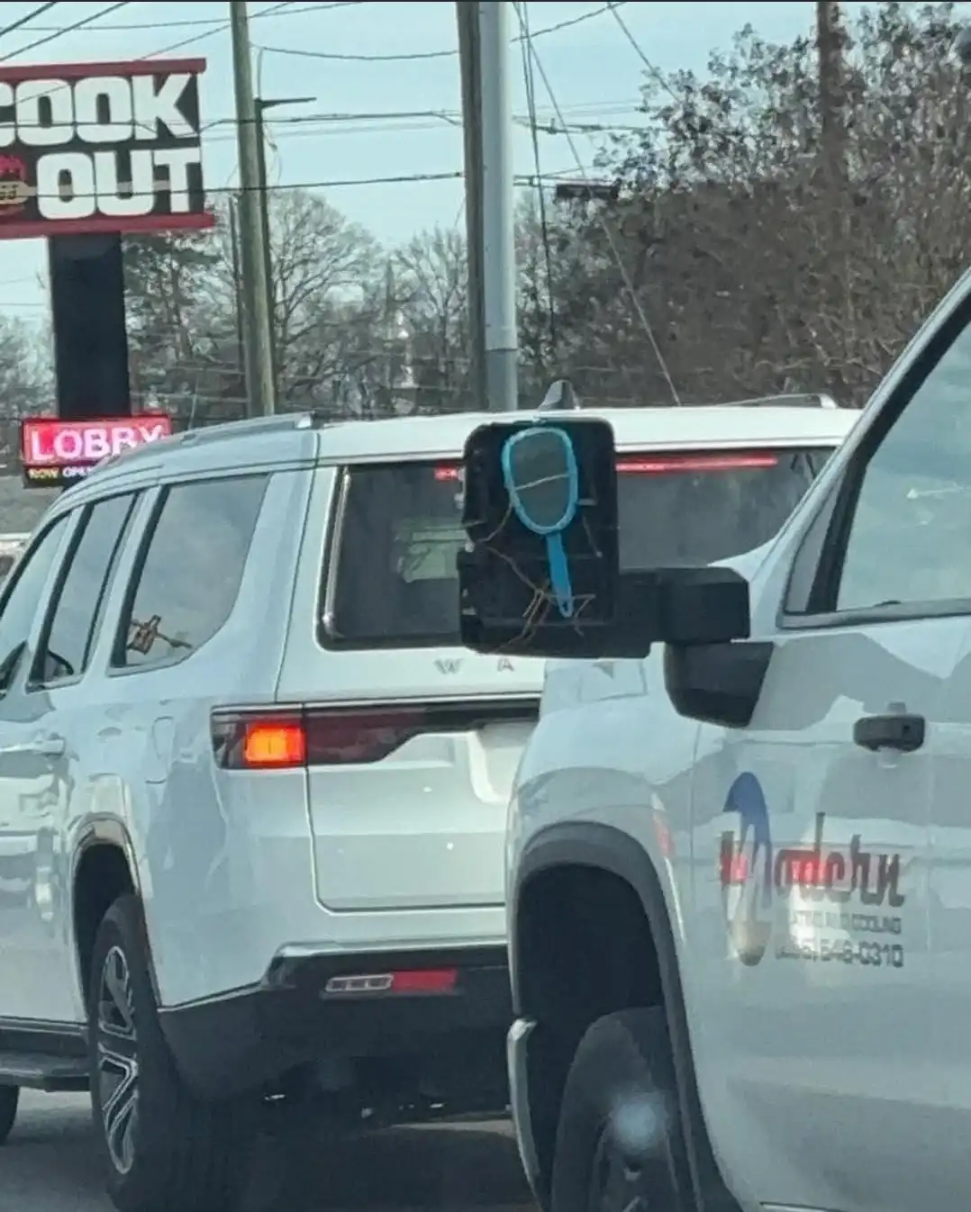A white SUV is stopped in traffic with a small, blue-handled hand mirror taped to its driver-side rearview mirror. In the…