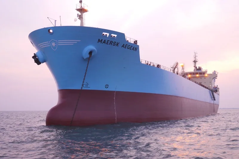 A large blue and red cargo ship named 'MAERSK AEGEAN' is shown at anchor in calm waters during sunset or sunrise, with its…
