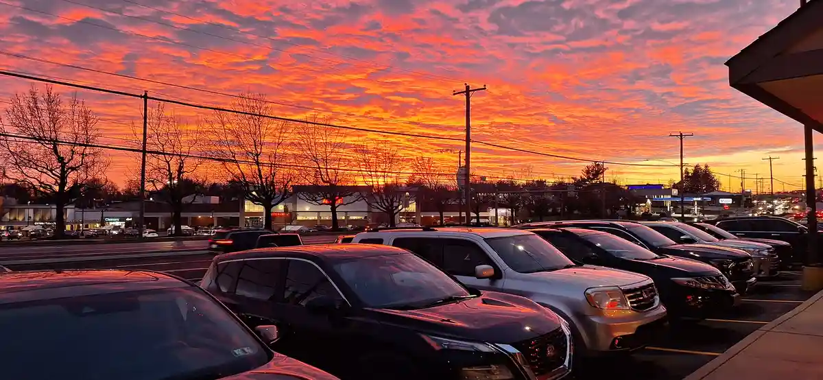 A parking lot filled with cars under a dramatic, fiery orange and red sunset sky, with bare trees and commercial buildings…