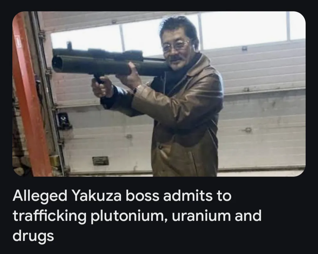 A man in a brown jacket and glasses holds a large, dark-colored rocket launcher in a garage-like setting with a white…