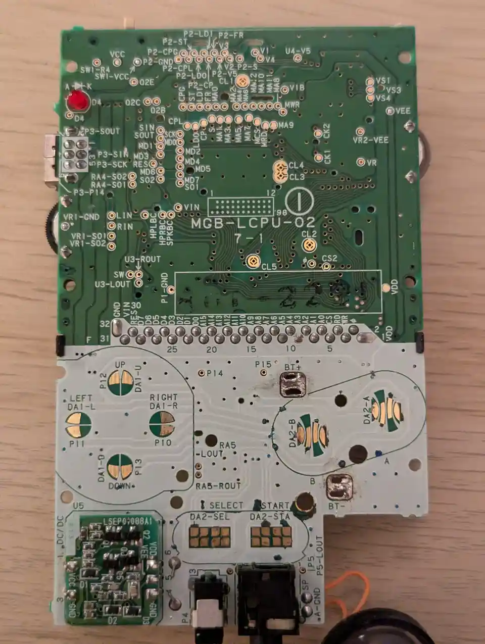 A close-up photo of a disassembled green and white circuit board, likely from a gaming console, showing exposed solder…