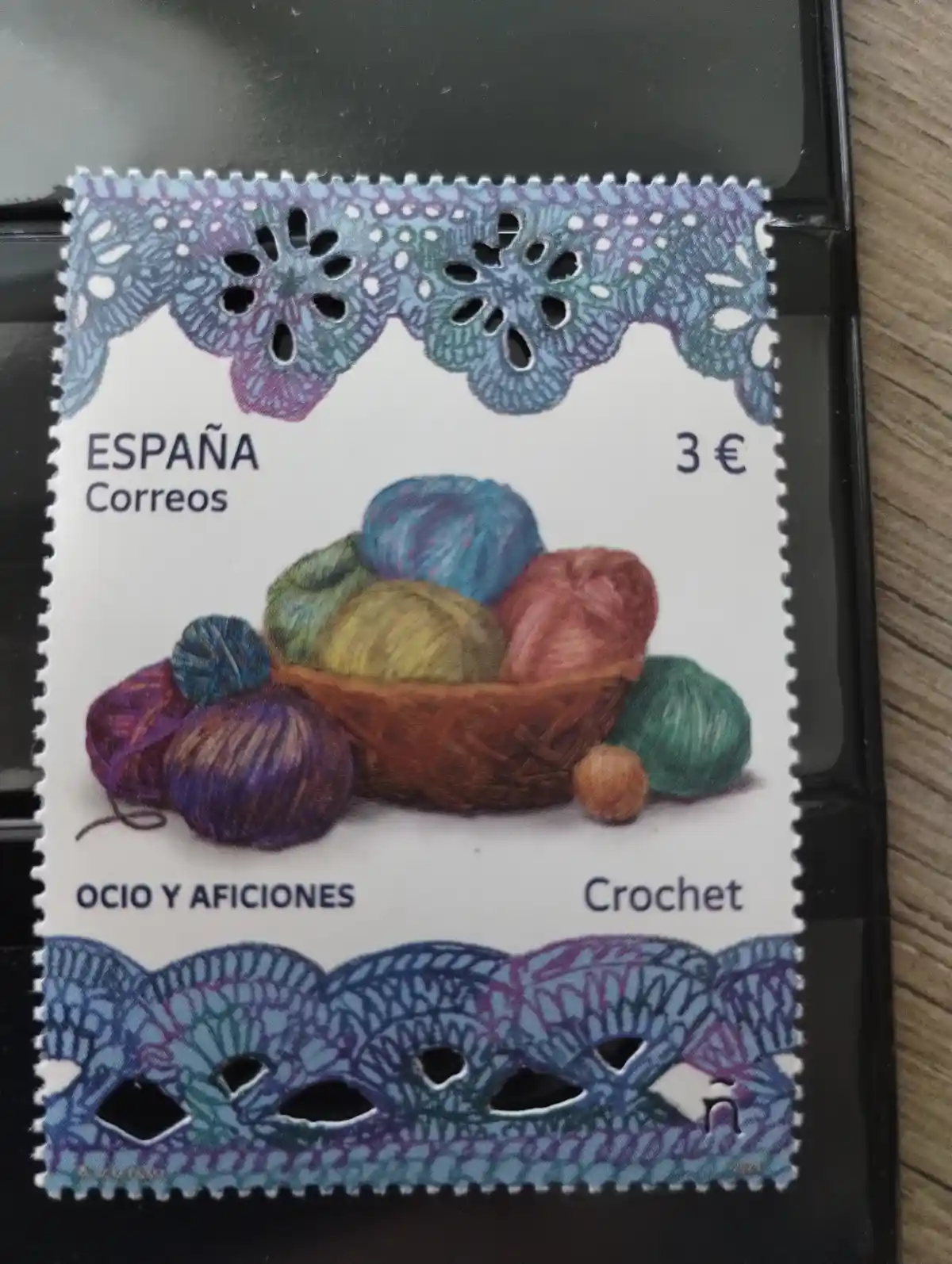 A Spanish postage stamp from Correos featuring a basket of colorful yarn balls and decorative crocheted lace borders. The…
