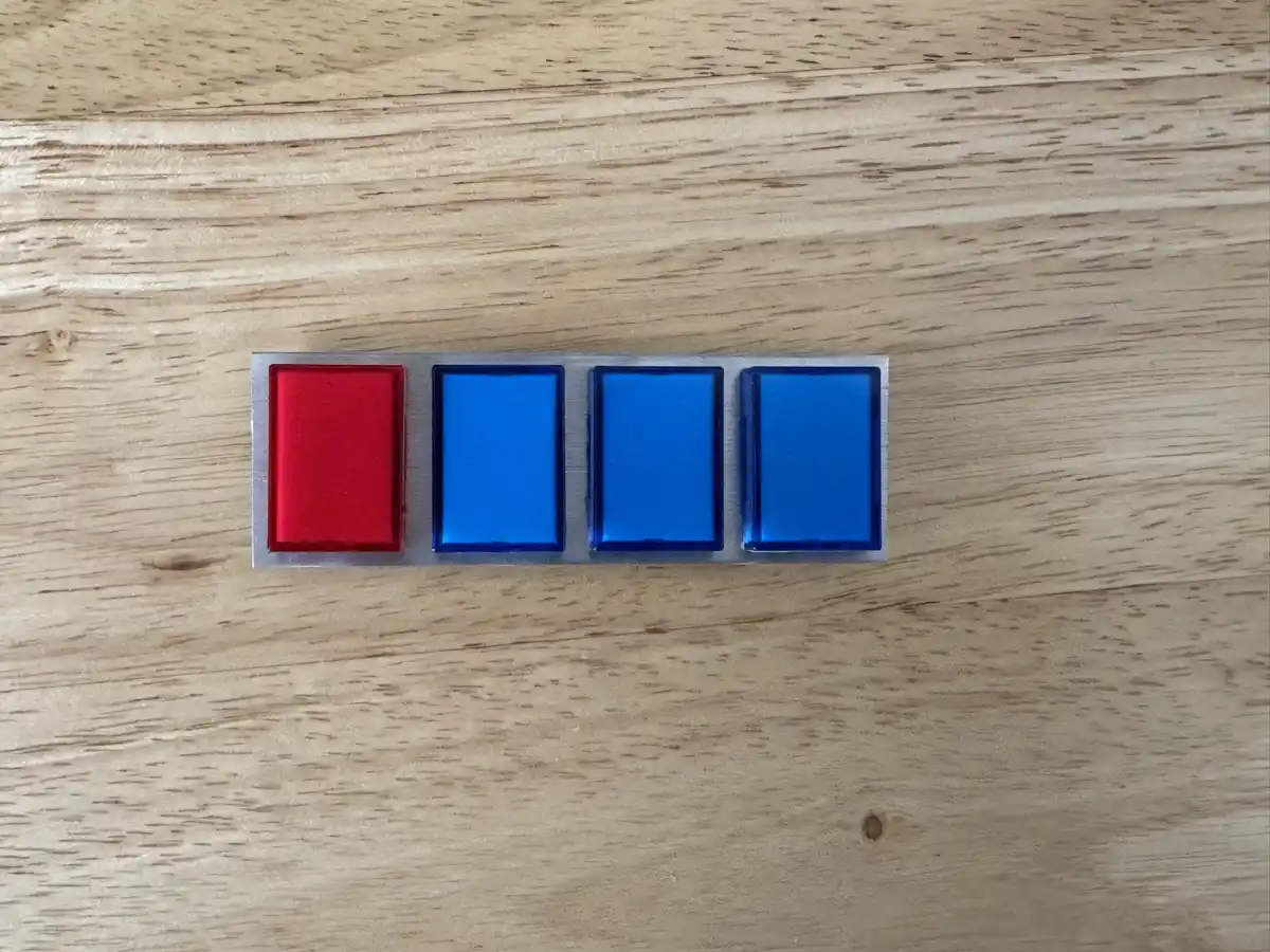 A close-up photo shows four rectangular buttons in a row on a wooden surface, with the leftmost button being red and the…