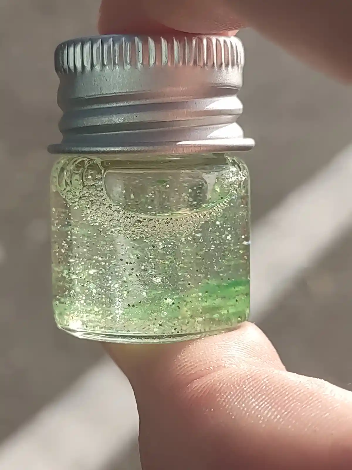 A close-up photo shows a hand holding a small, clear glass jar with a silver screw-top lid. Inside the jar is a green…