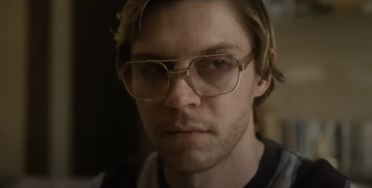 A close-up shot of a man with light brown hair and large, gold-rimmed glasses, looking intensely at the camera with a…