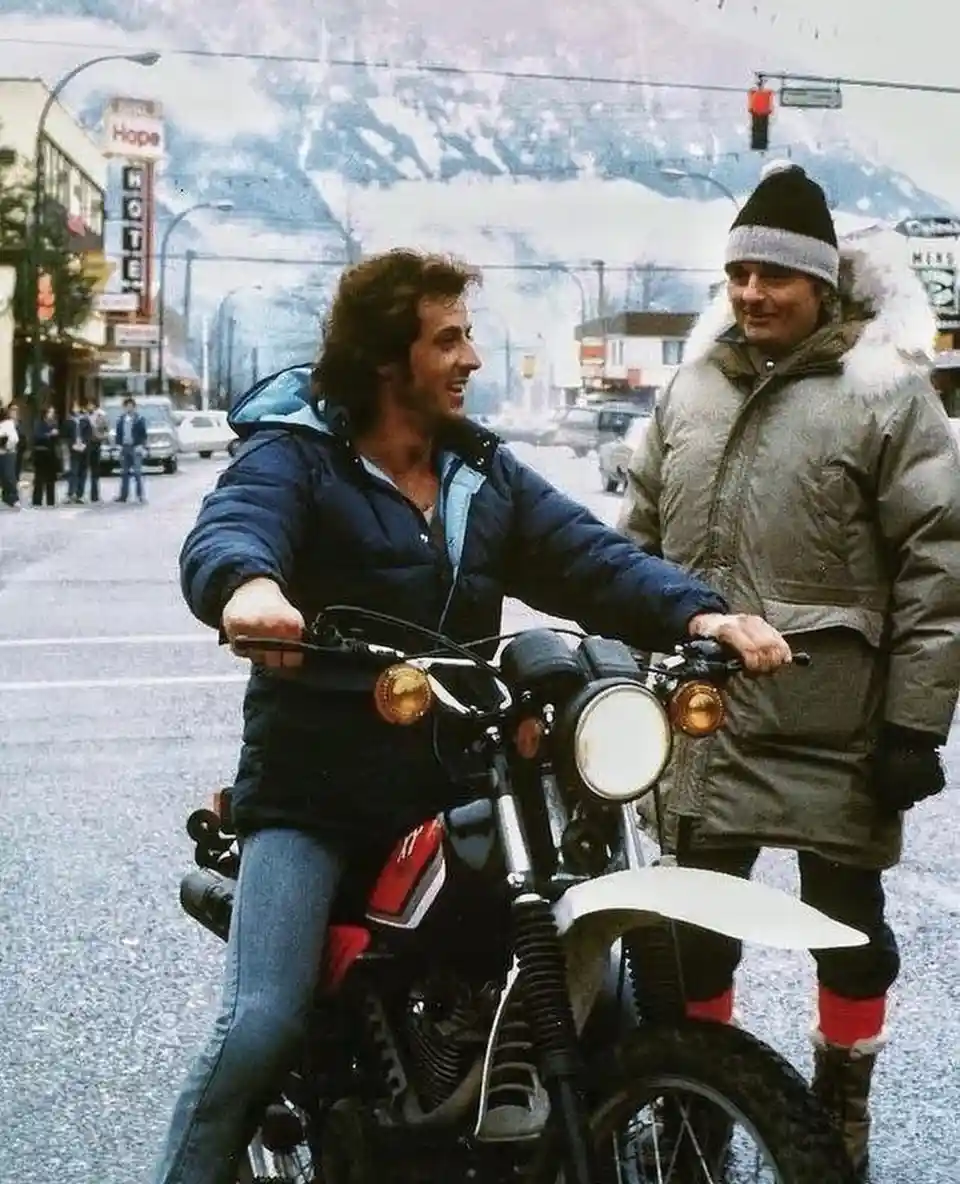 A man in a blue puffer jacket sits on a motorcycle, smiling at another man standing beside him wearing a thick winter coat…