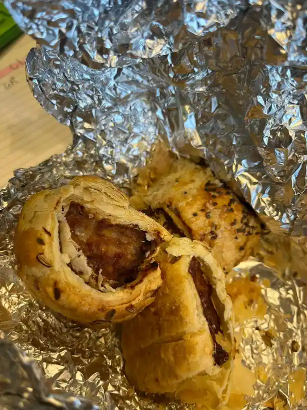 A close-up photo shows several golden-brown, flaky sausage rolls partially unwrapped from crinkled aluminum foil, with one…