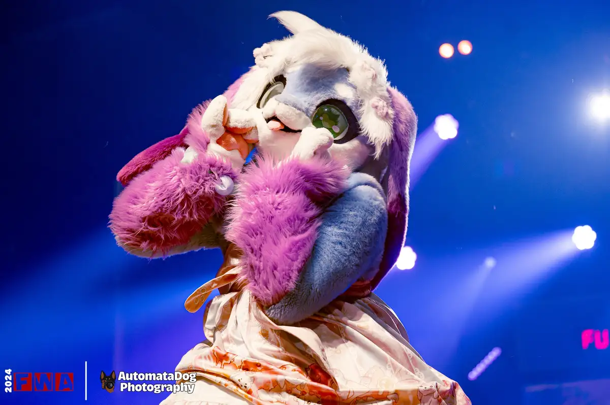 A person in a detailed blue and white fursuit with purple fluffy accents and large green eyes is on a stage with blue…