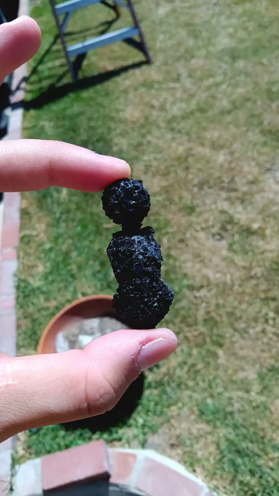 A hand holds a small, black, glossy object shaped vaguely like a human figure with a rose-like head, against a blurred…