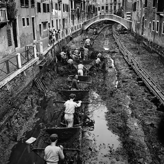 A black-and-white historical photo shows workers in Venice manually shoveling mud from a canal using small boats. The…