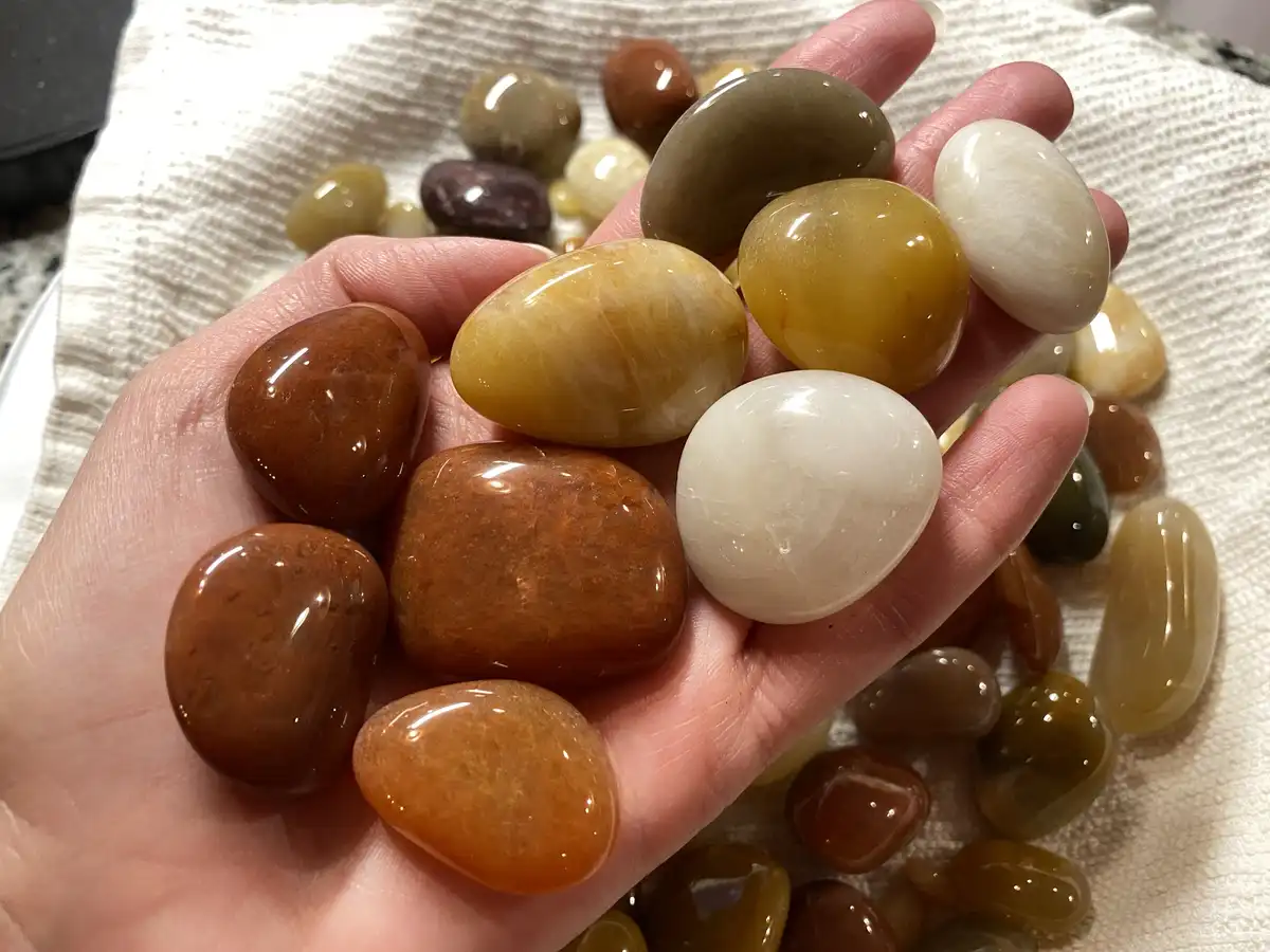 A person's hand holds a collection of smooth, polished, multi-colored stones resembling tumbled crystals or river rocks…