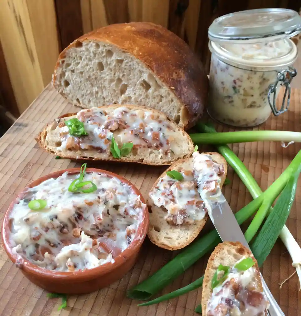 A rustic food photo shows a sliced loaf of bread with a creamy, chunky spread (possibly pâté or cheese dip) on slices…