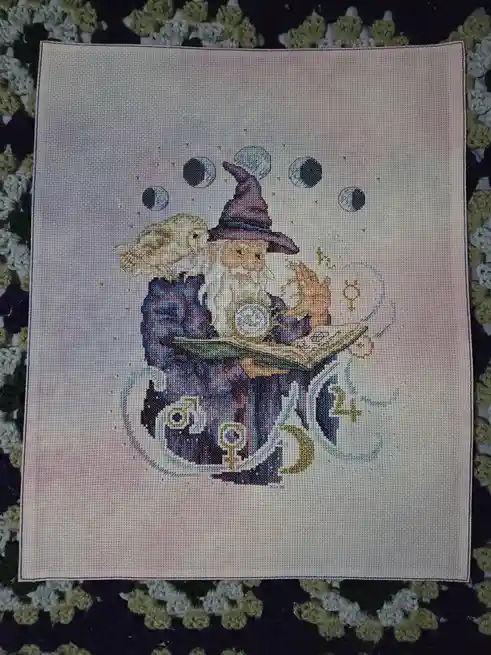 A cross-stitch embroidery depicts a wizard with a long white beard and purple hat, holding an open book, with an owl perched…