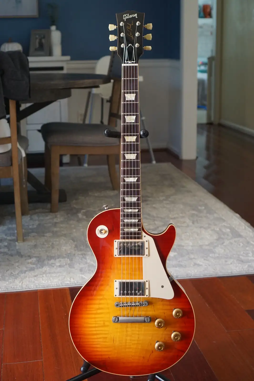A Gibson Les Paul electric guitar with a sunburst finish stands upright on a stand in a home setting, positioned on a…