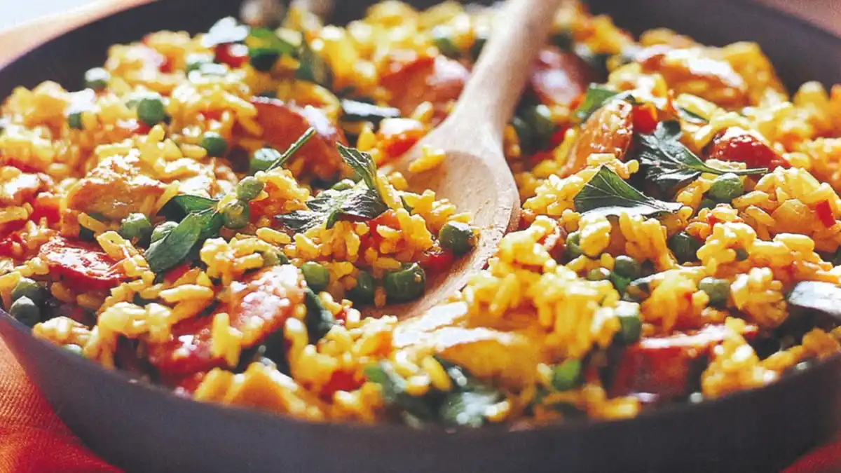 A close-up photo of a colorful rice dish in a black pan, featuring yellow rice mixed with green peas, red bell peppers…