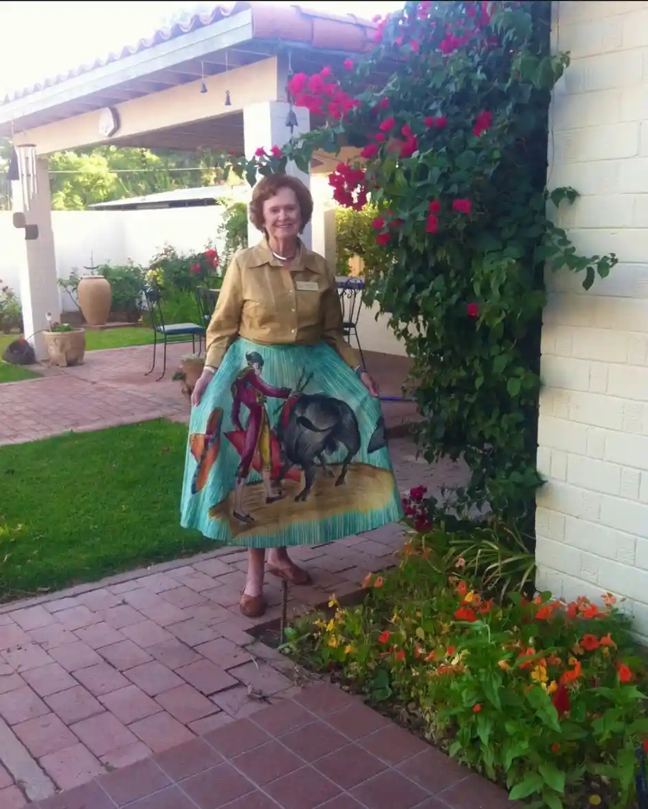 An elderly woman stands outdoors in a garden, smiling at the camera. She wears a tan blouse and a long, light blue skirt…