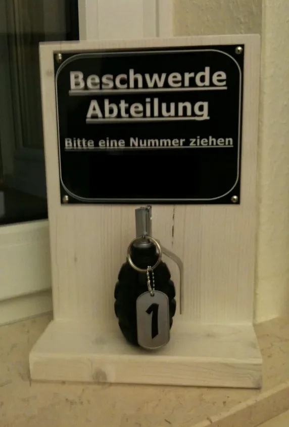 A wooden stand holds a black sign reading 'Beschwerde Abteilung' and 'Bitte eine Nummer ziehen', with a grenade-shaped…
