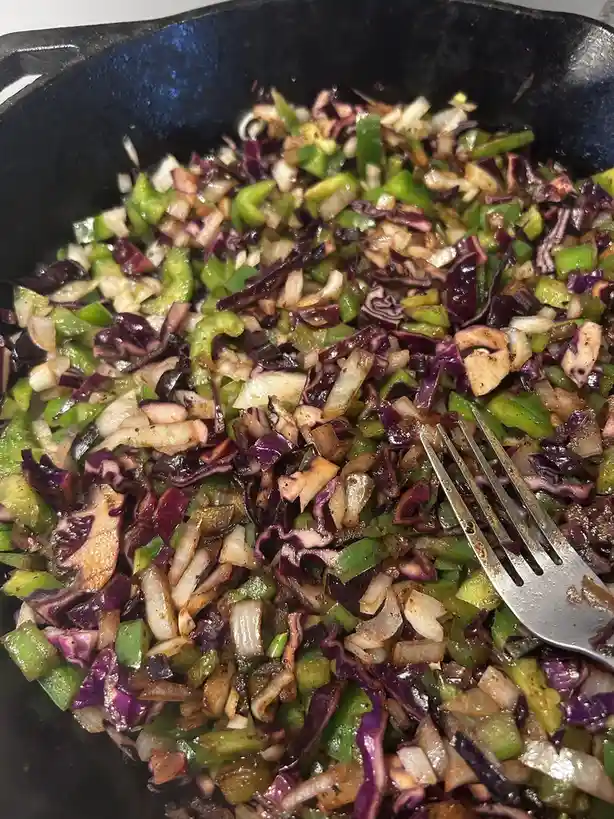A close-up photo shows a black cast-iron skillet filled with sautéed chopped vegetables, including green bell peppers, white…