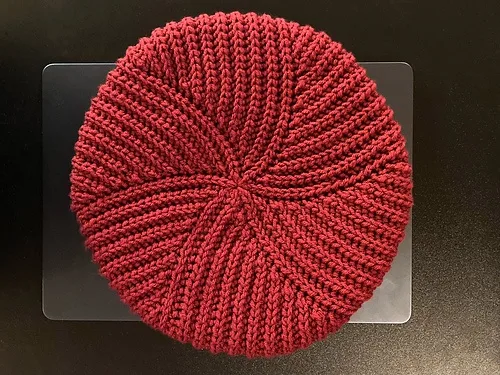 a red knitted beanie hat sits centered on a clear plastic tray against a dark background. The hat has a textured, ribbed…