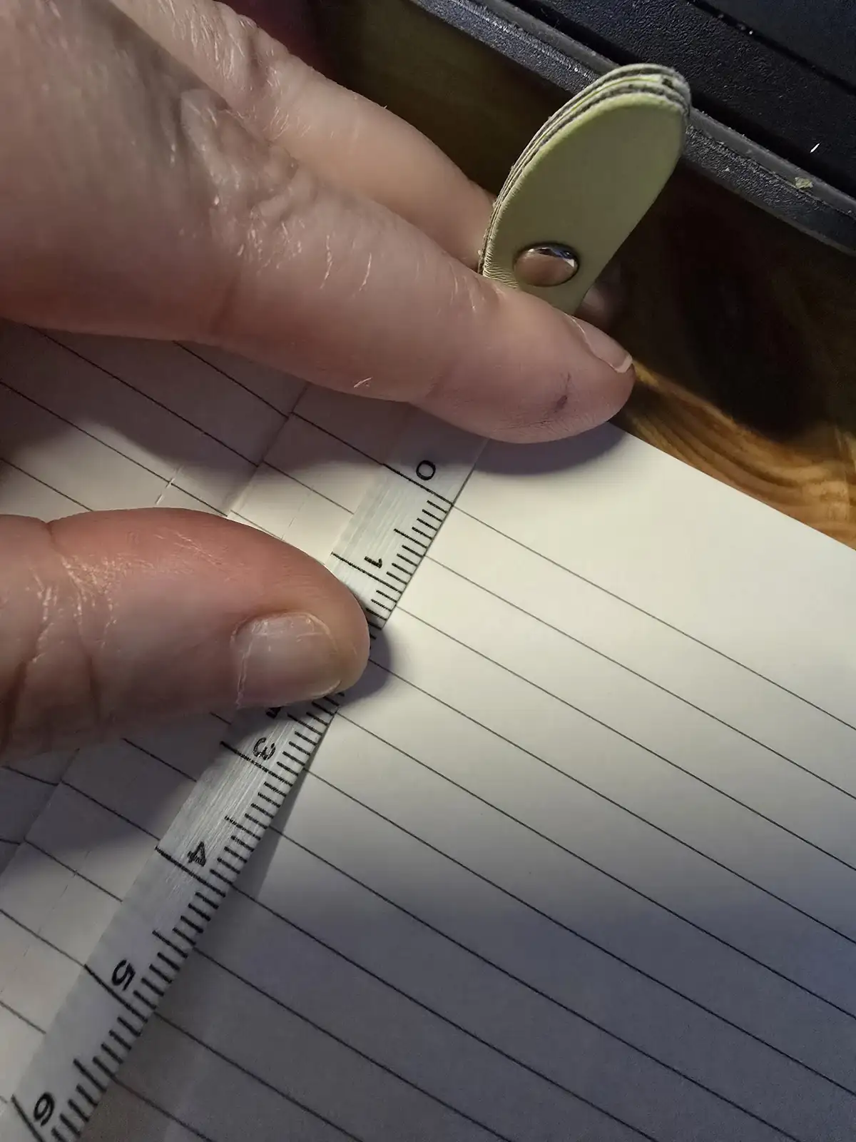 A close-up photo shows a person's fingers holding a small, light green leather object against a ruler placed on lined paper…