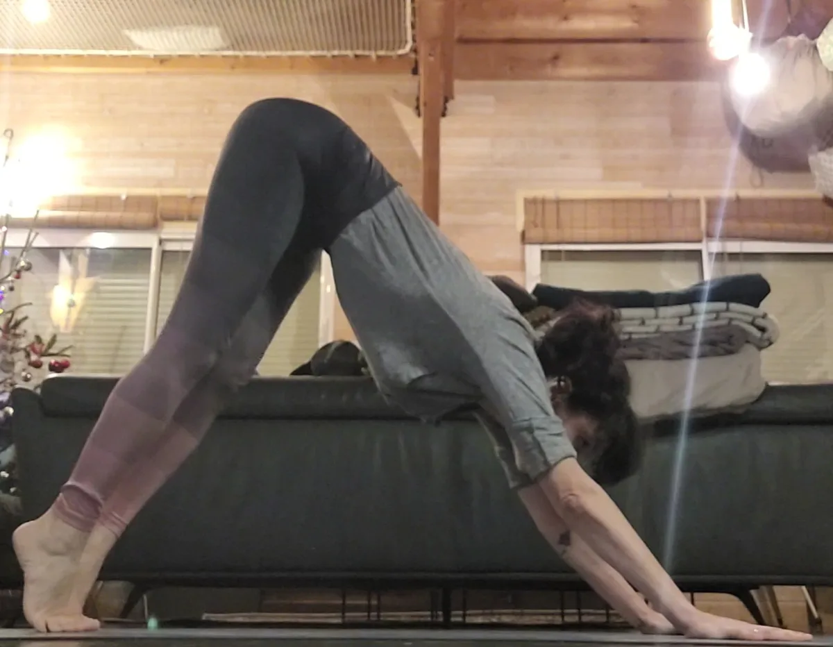 a person in leggings and a grey shirt is doing a yoga pose with their head down on a dark couch, arms extended forward, feet…