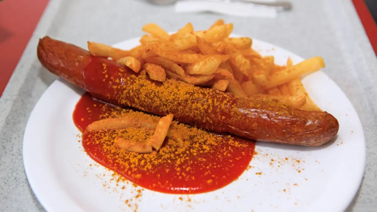 A close-up photo shows a sausage on a white plate with french fries, both generously dusted with orange spice powder and…