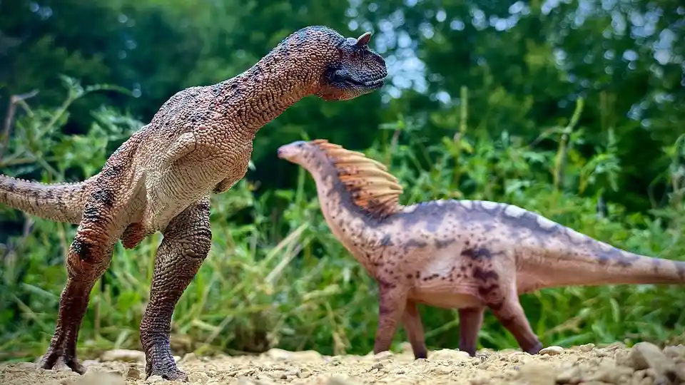 Two detailed dinosaur figurines stand in a grassy outdoor setting. The foreground dinosaur, resembling a ceratopsian with a…