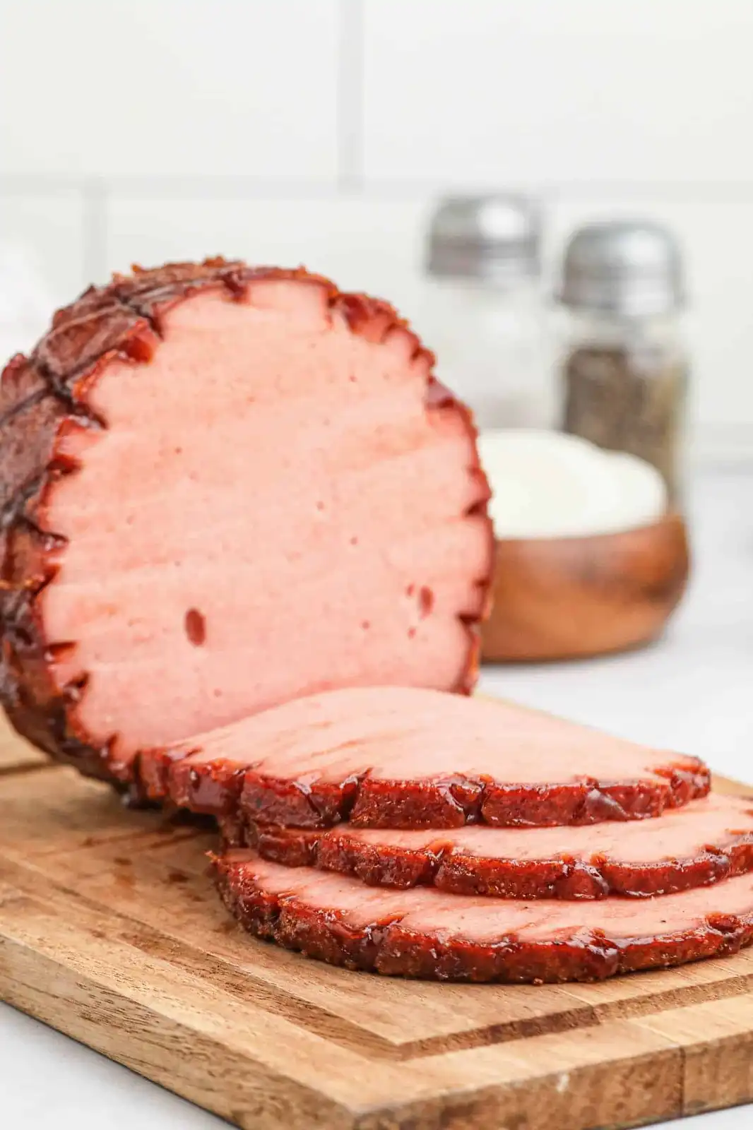 A close-up photo shows a glazed, sliced ham on a wooden cutting board, with a few slices fanned out in front. In the blurred…