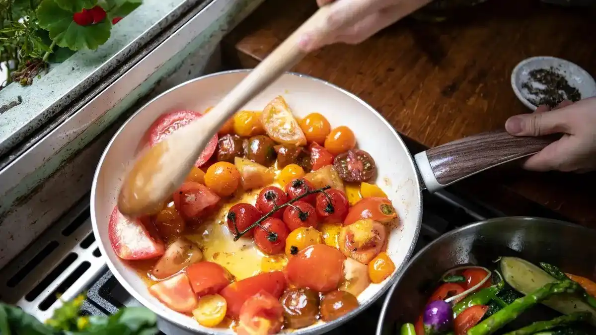 A close-up shot shows hands stirring a white frying pan filled with various colorful tomatoes—red, yellow, and…