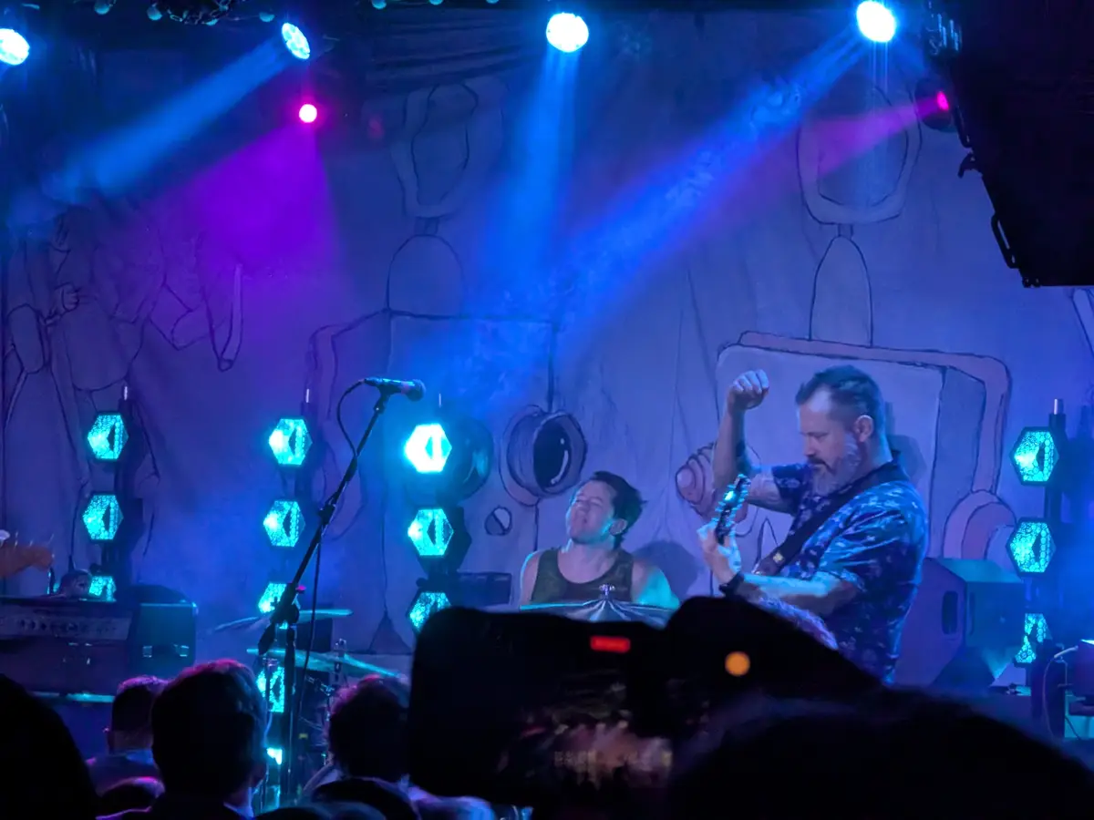 A live concert photo shows two musicians on stage under blue and purple lights, with a cartoonish backdrop. The guitarist on…