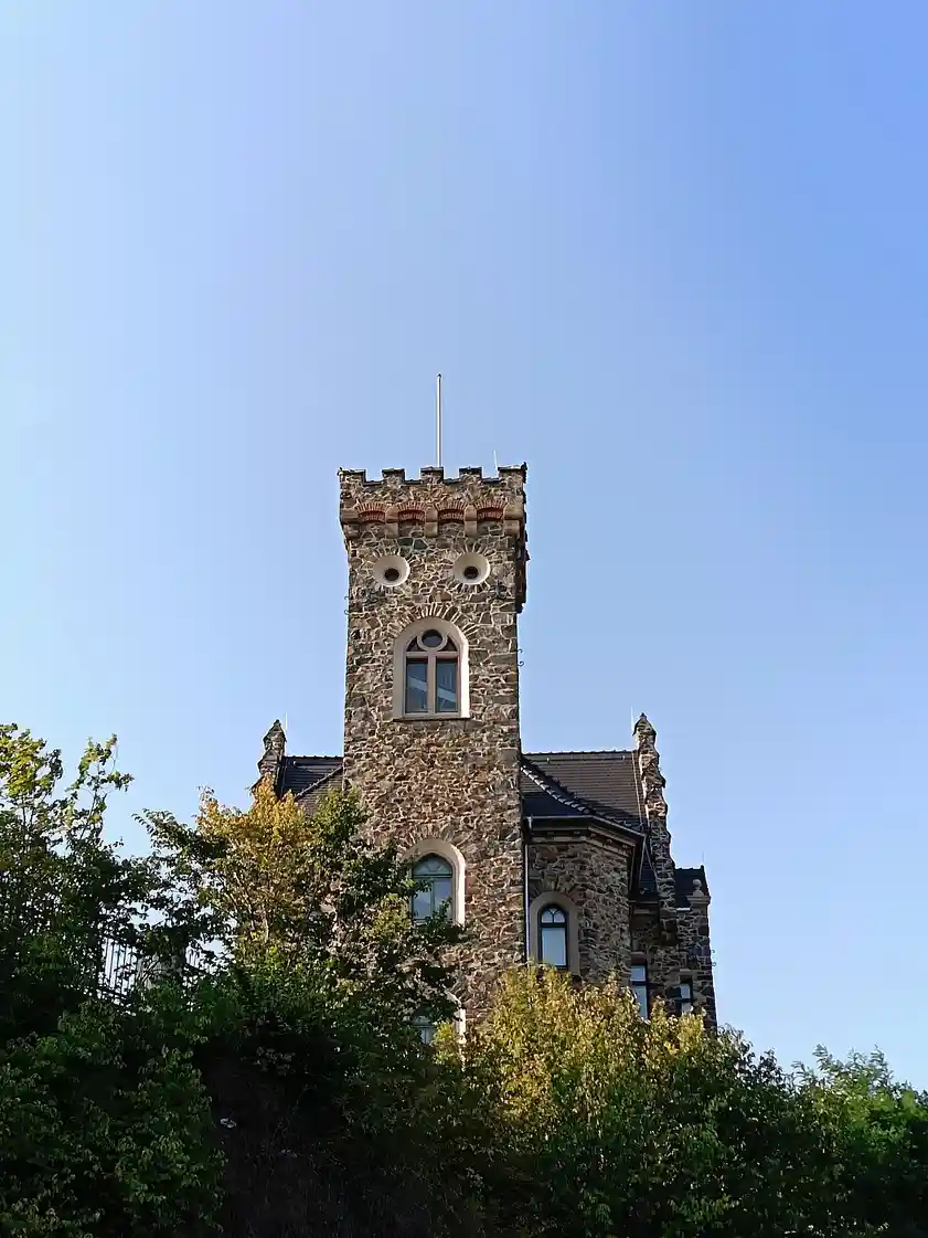 A stone castle tower with arched windows and crenellations stands against a clear blue sky, partially obscured by green…