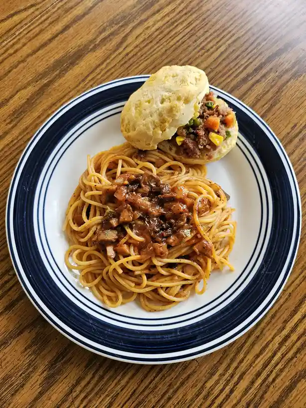 A plate of spaghetti with meat sauce sits next to a biscuit filled with meat and vegetables, both on a white plate with blue…