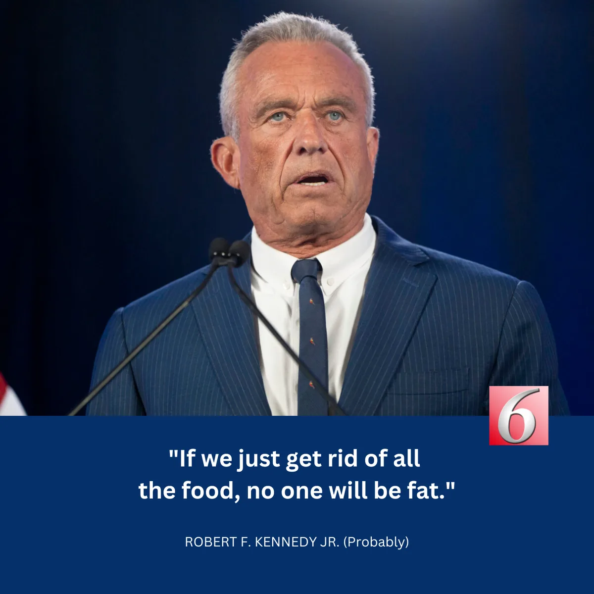 A man with gray hair and blue eyes, wearing a blue pinstripe suit and tie, stands at a podium with microphones against a…