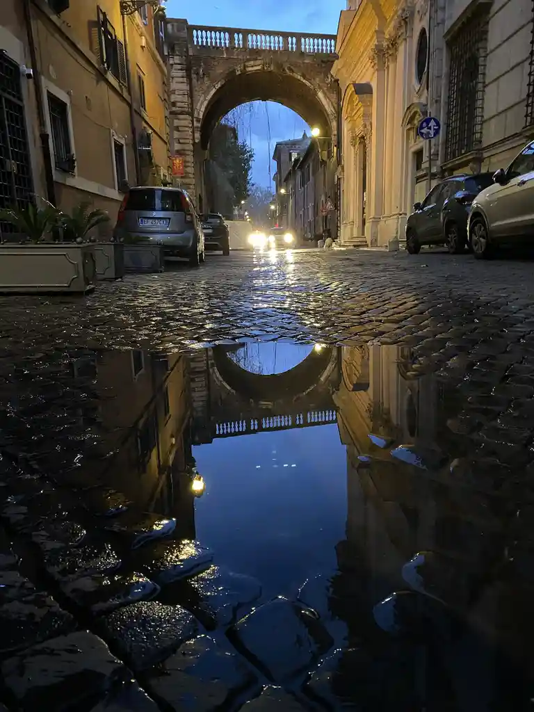 A cobblestone street at dusk with a large puddle reflecting an arched stone bridge and surrounding buildings. Cars are…