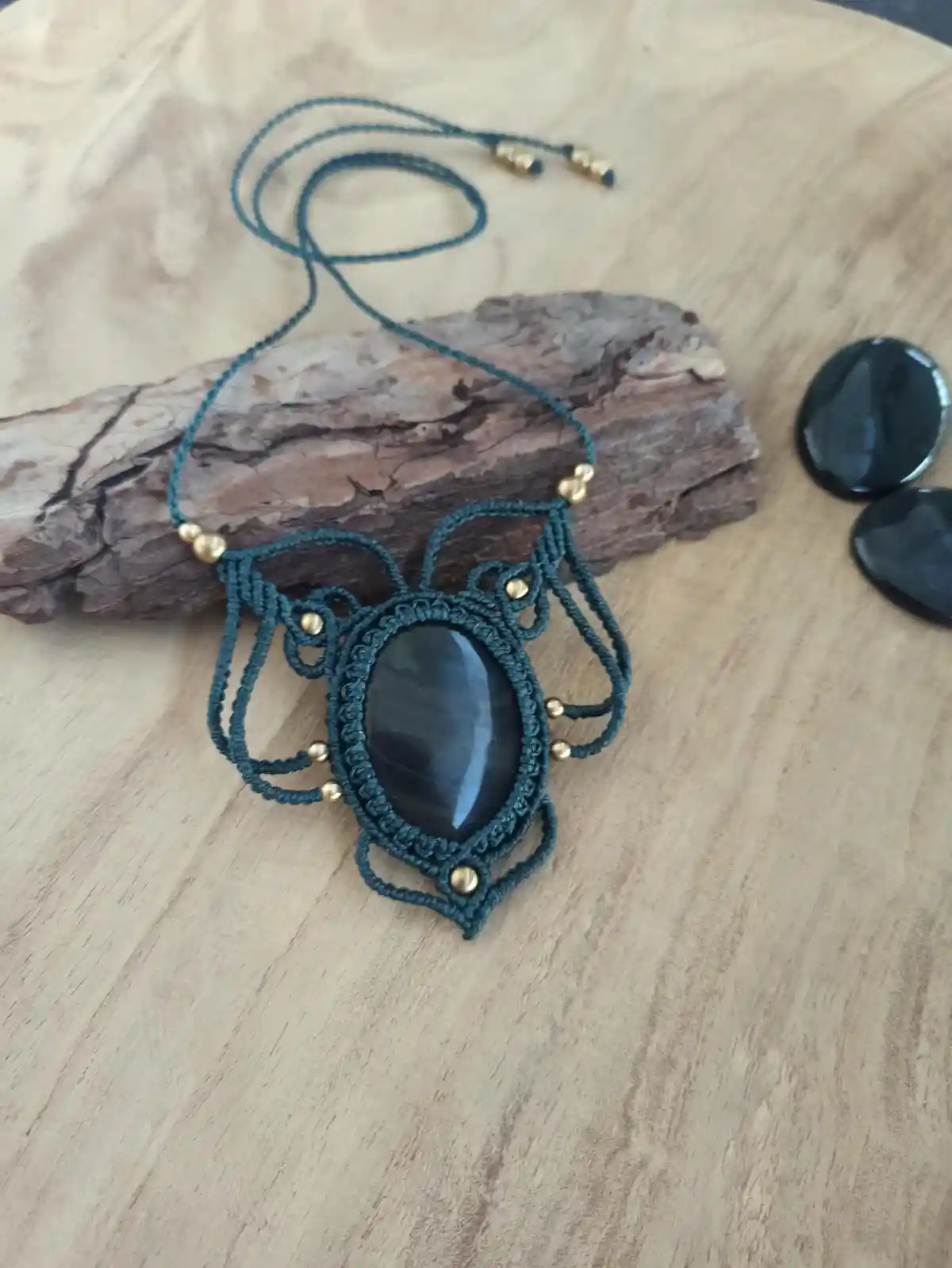 A handmade macrame necklace with a dark blue cord, gold beads, and a central black oval stone pendant rests on a piece of…