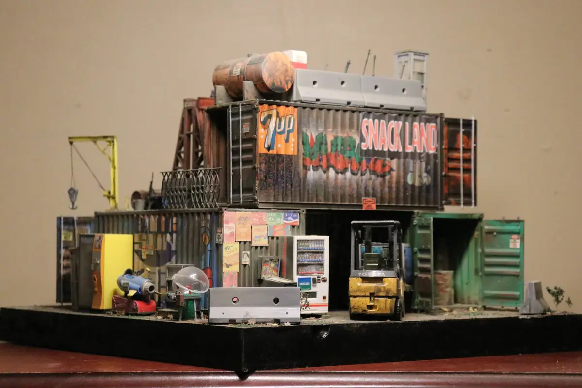 A detailed miniature diorama of a cluttered, multi-level structure built from stacked shipping containers, featuring a '7up'…