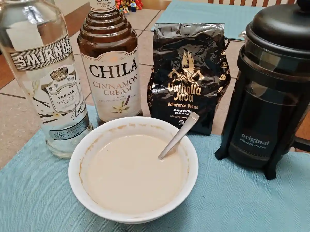 A white bowl containing a beige liquid sits on a blue placemat, surrounded by a bottle of Smirnoff Vanilla vodka, a bottle…