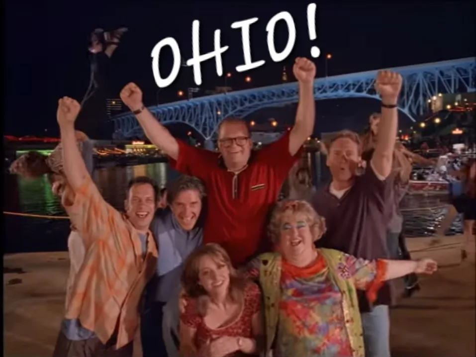 A group of people, including a man in a red shirt and glasses, are cheering with raised fists at night near a lit bridge…