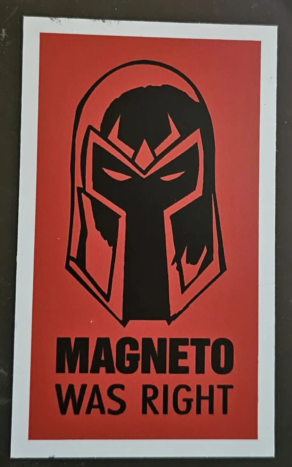 A red sticker features a black silhouette of Magneto's iconic helmet with angular, menacing design. Below the helmet, bold…