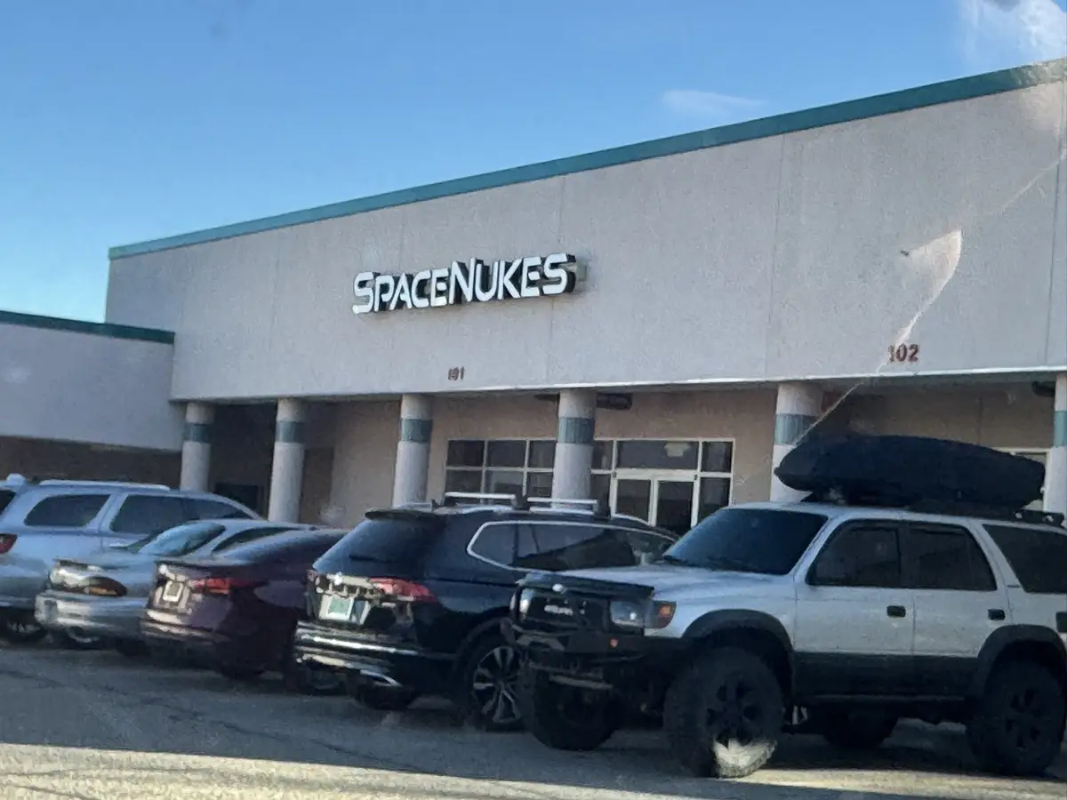 An exterior shot of a commercial building with a sign that reads 'SPACE NUKES' above the entrance, with several cars parked…