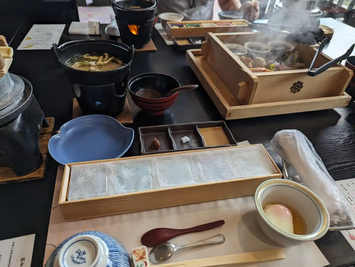 a table laden with traditional japanese dining items including a steaming pot, a wooden box with food, a bowl of soup, a…