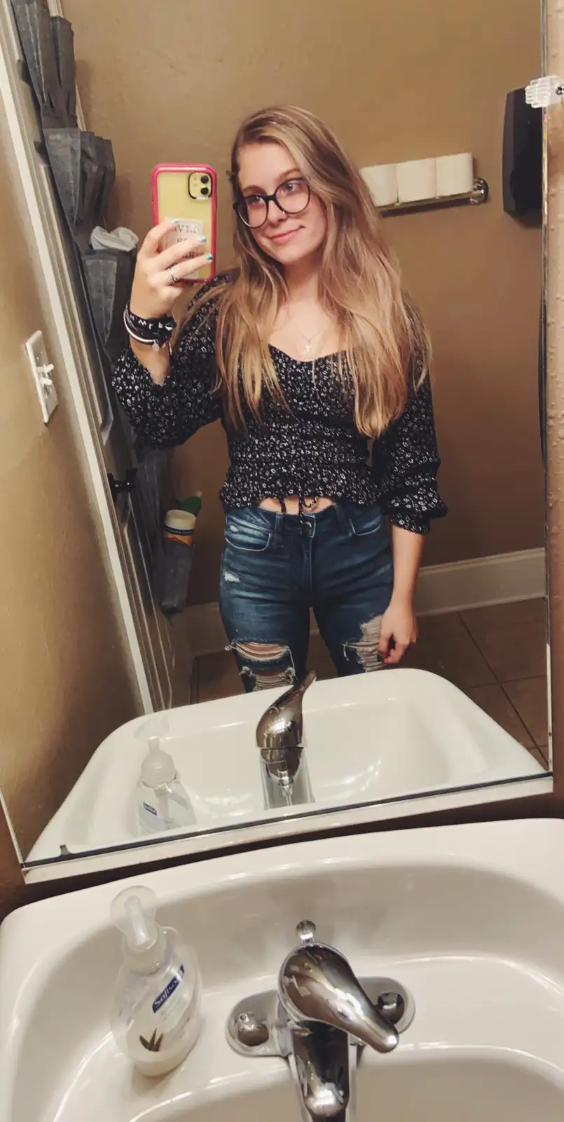 A young woman with long blonde hair and glasses takes a mirror selfie in a bathroom, holding a pink-cased phone. She wears a…