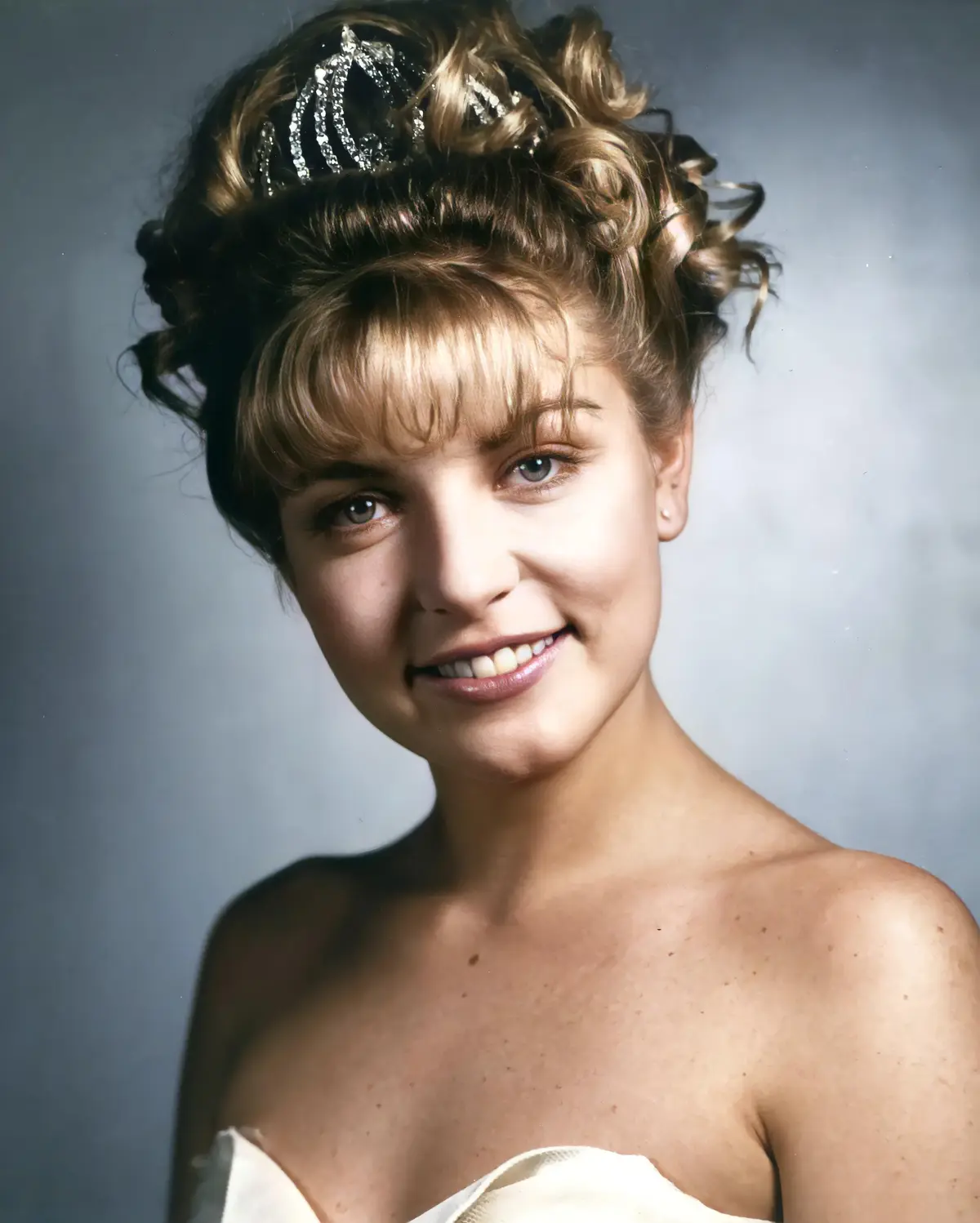 A young woman with styled blonde hair and a sparkling tiara smiles at the camera, wearing a strapless white dress against a…