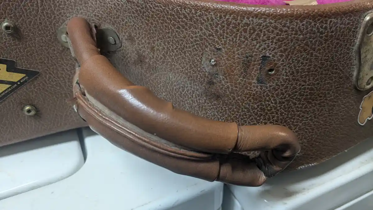 A close-up of a worn, brown leather suitcase handle that is visibly frayed, torn, and bent at the end, resting on a white…