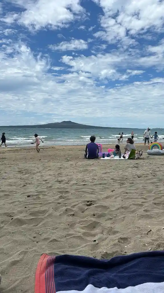 A wide beach scene with people relaxing and playing near the water under a partly cloudy sky. In the foreground, a towel is…